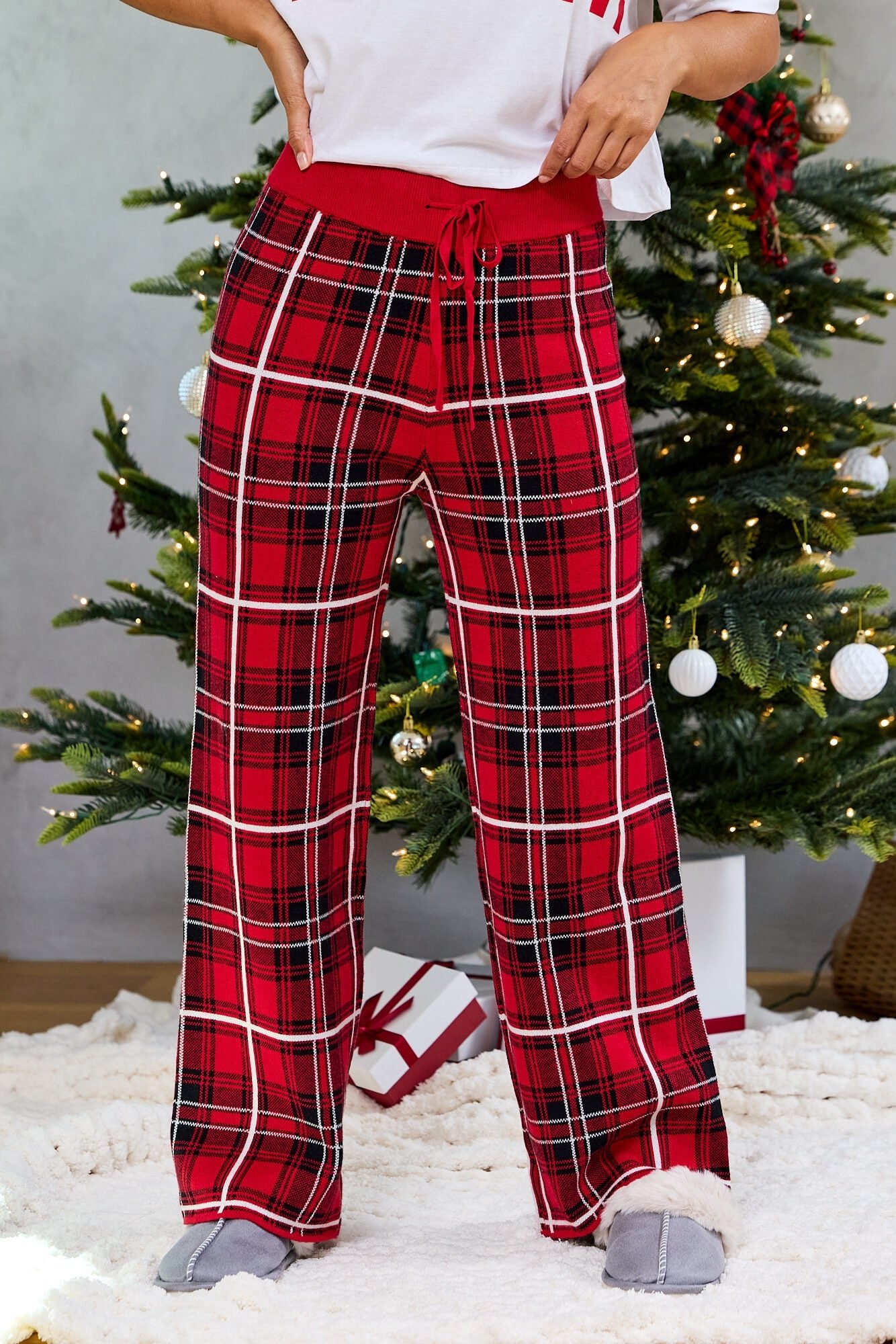 Wearing Fireside Red Plaid Sweater Pajama Pants and slippers, a person stands by a decorated Christmas tree with gifts underneath.