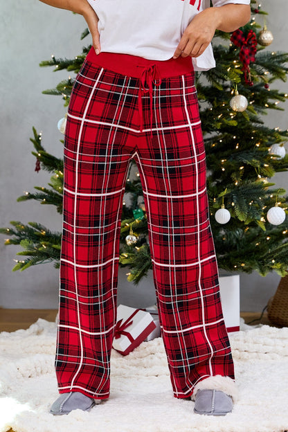 Wearing Fireside Red Plaid Sweater Pajama Pants and slippers, a person stands by a decorated Christmas tree with gifts underneath.