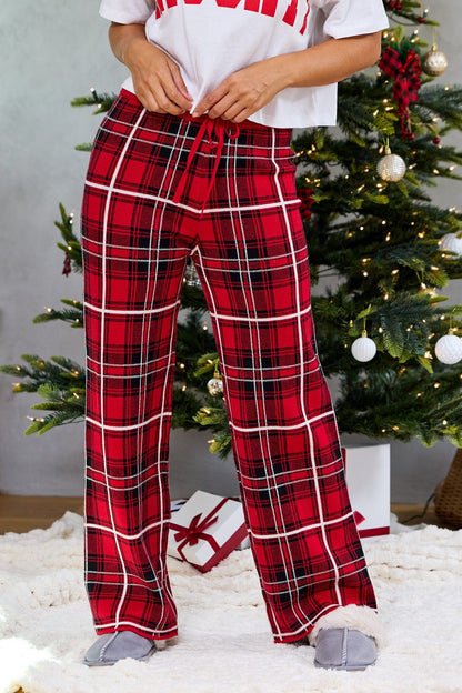Person in Fireside Red Plaid Sweater Pajama Pants and slippers stands by a decorated Christmas tree with gifts underneath.