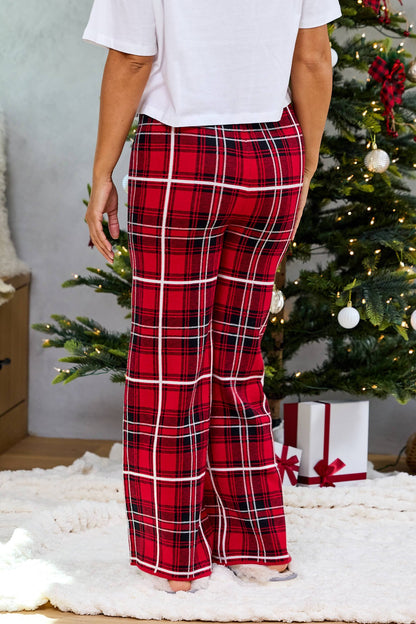 Person in Fireside Red Plaid Sweater Pajama Pants stands by a decorated Christmas tree with wrapped gifts on a white rug.