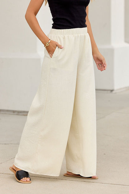 Woman in First Class Wide Leg Pants - Natural, black sleeveless top, sandals, gold bracelet, stylish on a city sidewalk.