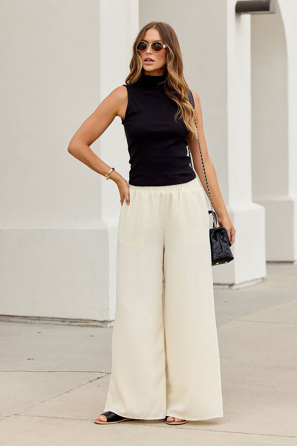 Woman in sunglasses and sleeveless black top wears First Class Wide Leg Pants - Natural, sandals, and holds a black purse outdoors.