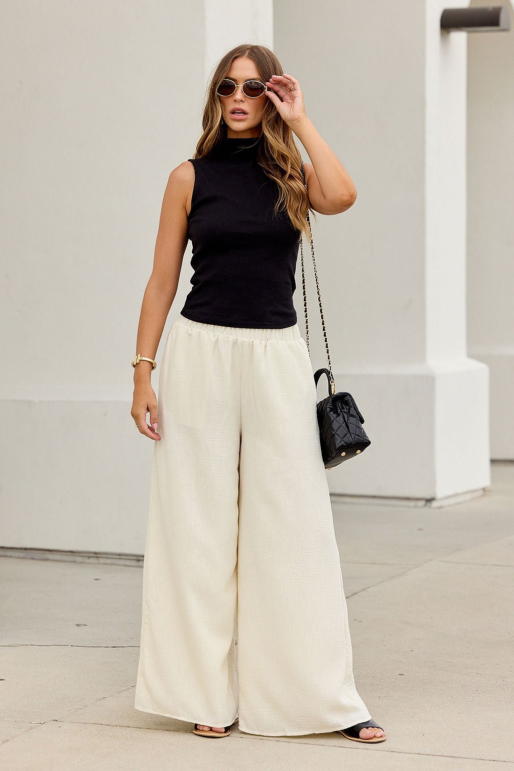 Woman in natural sunglasses, black sleeveless top, and First Class Wide Leg Pants - Natural stands on a sidewalk with a black purse.