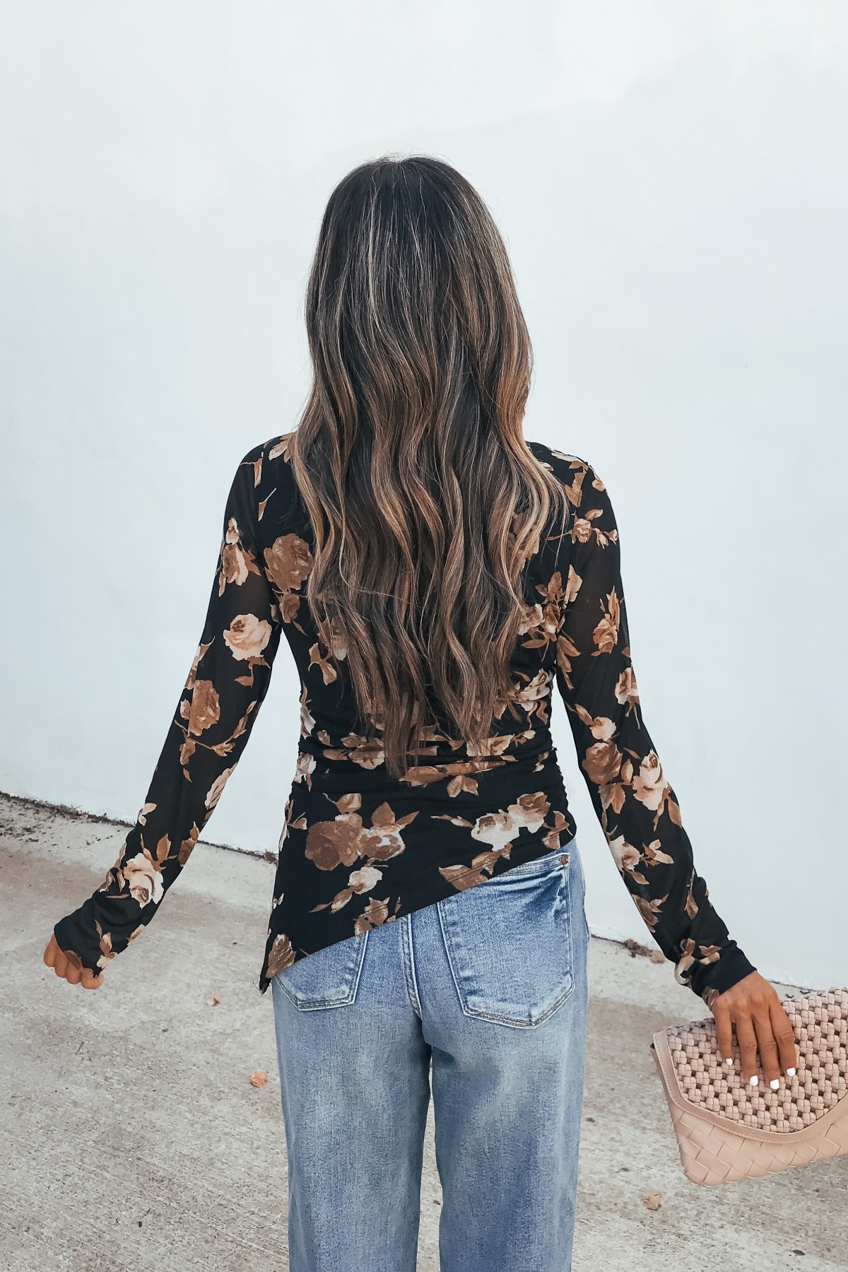 Woman with long wavy hair wears a black Floral Print Asymmetrical Top and jeans, facing away, holding a textured beige purse.