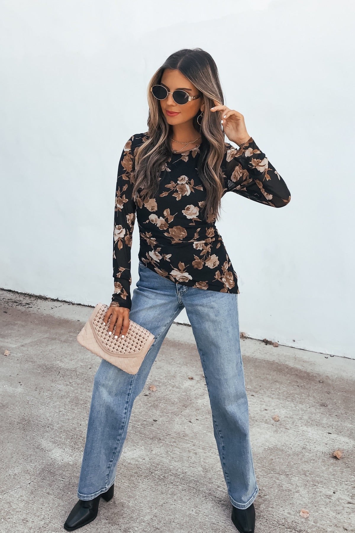 Woman in sunglasses, Floral Print Asymmetrical Top - Black, blue jeans, black boots, beige clutch stands by a white wall.