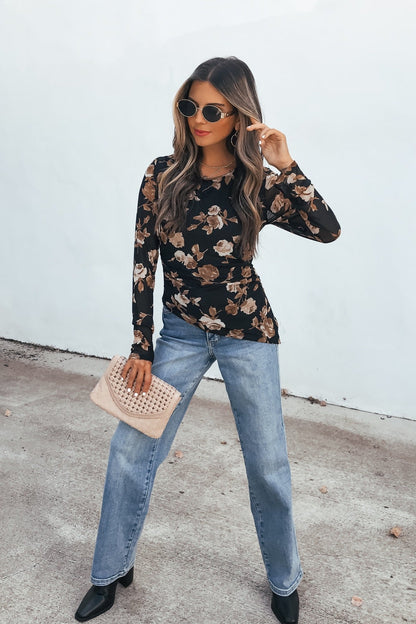 Woman in sunglasses, Floral Print Asymmetrical Top - Black, blue jeans, black boots, beige clutch stands by a white wall.