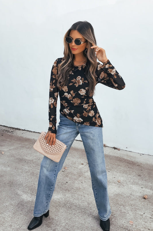 Woman in sunglasses, Floral Print Asymmetrical Top - Black, blue jeans, black boots, beige clutch stands by a white wall.