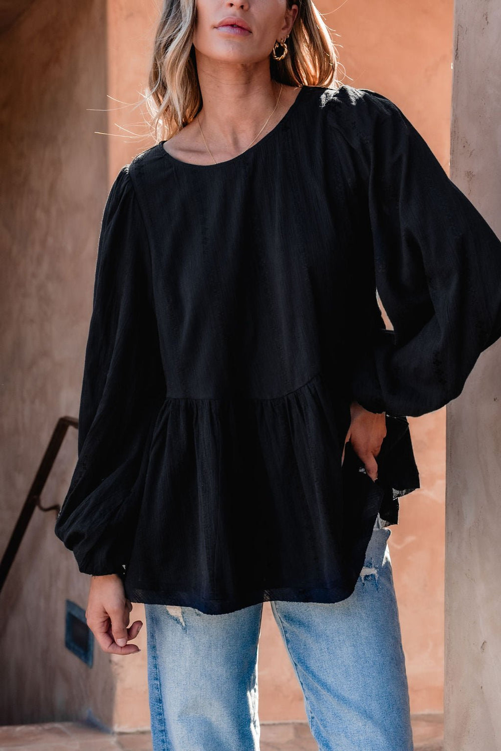 Woman in a flowy black ruffled tunic—DOORBUSTER—paired with ripped jeans, standing by a peach-colored wall.