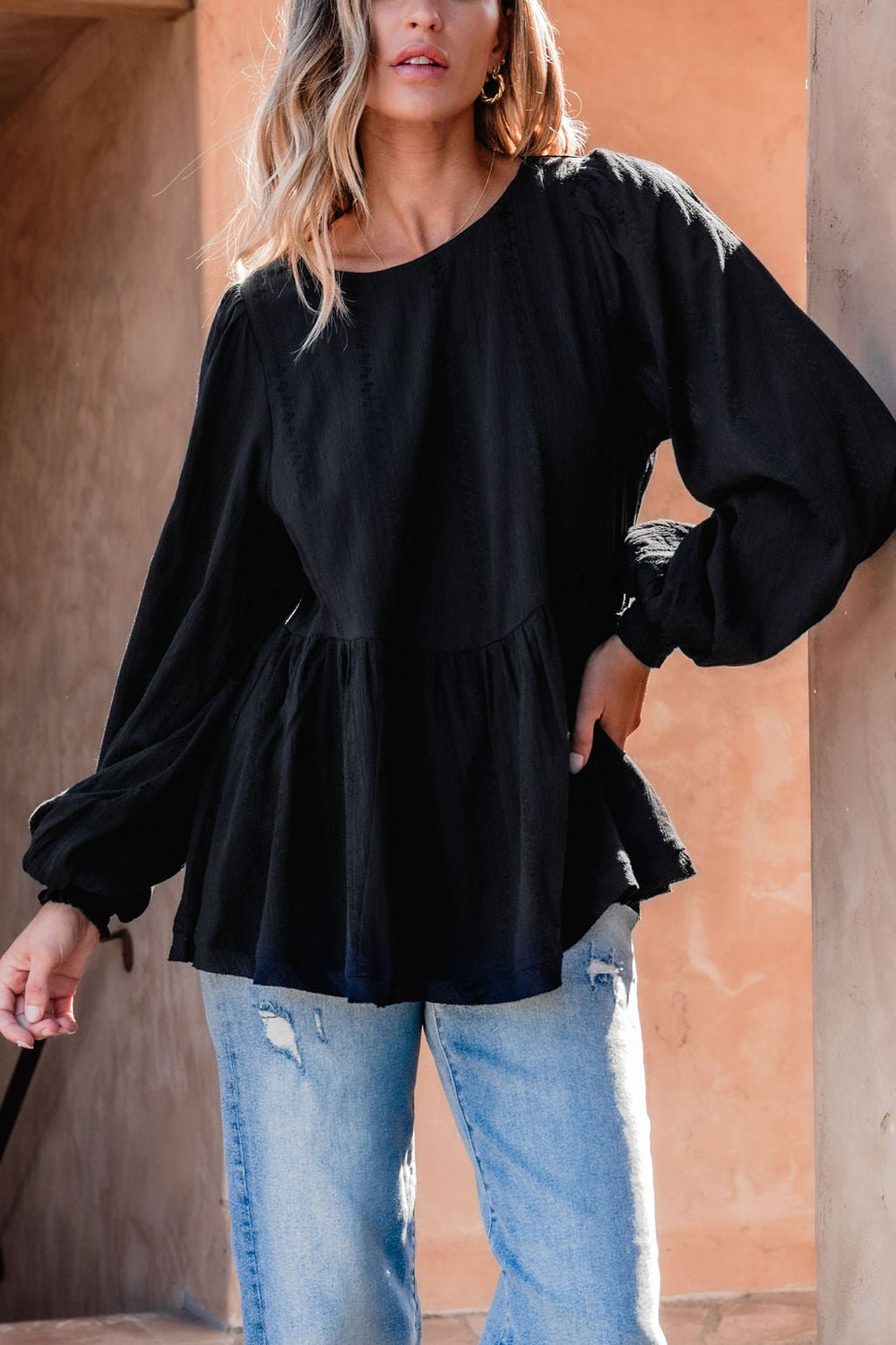 Woman in a Flowy Black Ruffled Tunic - DOORBUSTER, with light ripped jeans, stands by a tan textured wall.