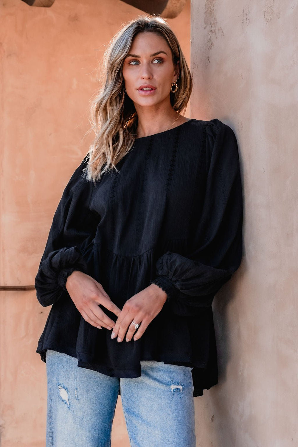 Woman with wavy blonde hair wears the Flowy Black Ruffled Tunic - DOORBUSTER and ripped blue jeans, standing by a beige wall.