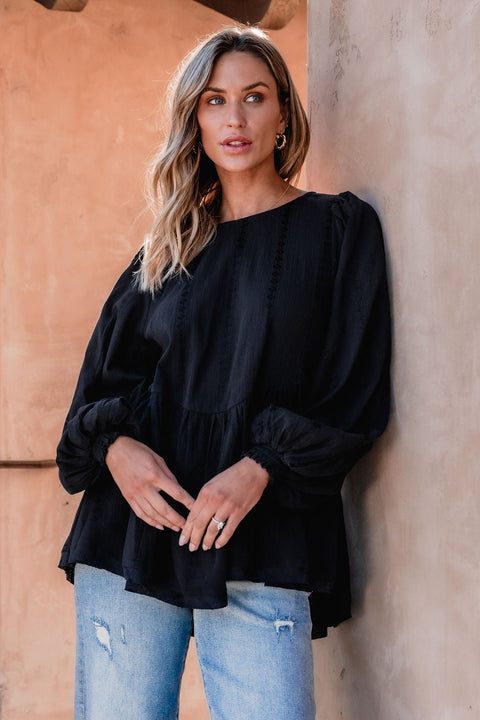 Woman with wavy blonde hair wears the Flowy Black Ruffled Tunic - DOORBUSTER and ripped blue jeans, standing by a beige wall.