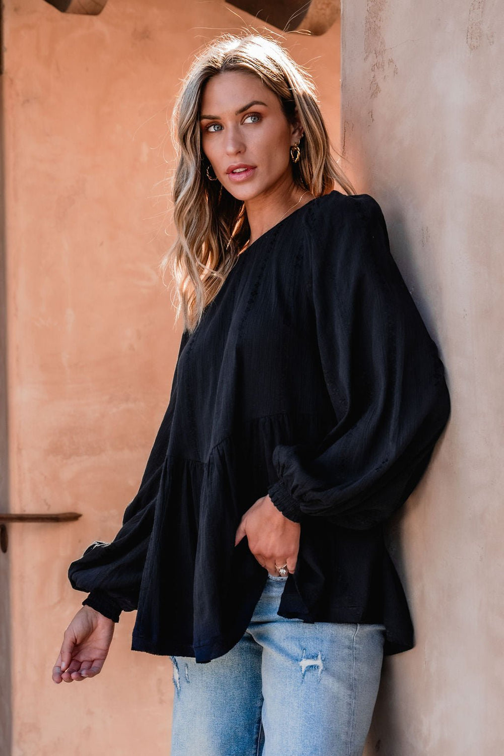 Woman with wavy blonde hair models the Flowy Black Ruffled Tunic - DOORBUSTER with light blue jeans against a tan textured wall.