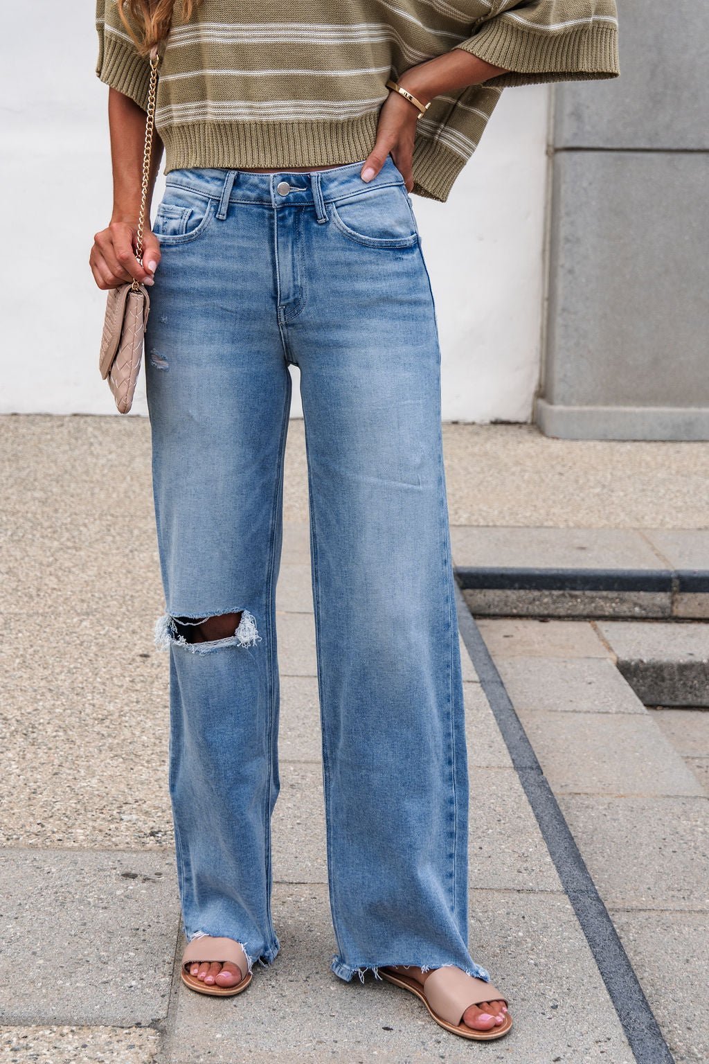 A person wears an olive-striped sweater, Flying Monkey Distressed Raw Hem Jeans - Light Wash, tan sandals, and holds a beige quilted purse.