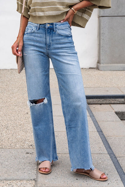 Person wears Flying Monkey Distressed Raw Hem Jeans - Light Wash, beige sandals, striped sweater, holding a small purse; only legs visible.