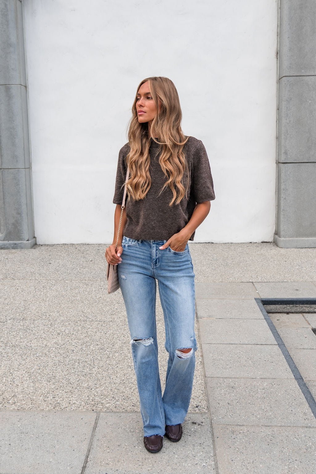 A woman with long wavy blonde hair stands outside in a brown sweater and Flying Monkey Distressed Wide Leg Jeans - Light Wash.