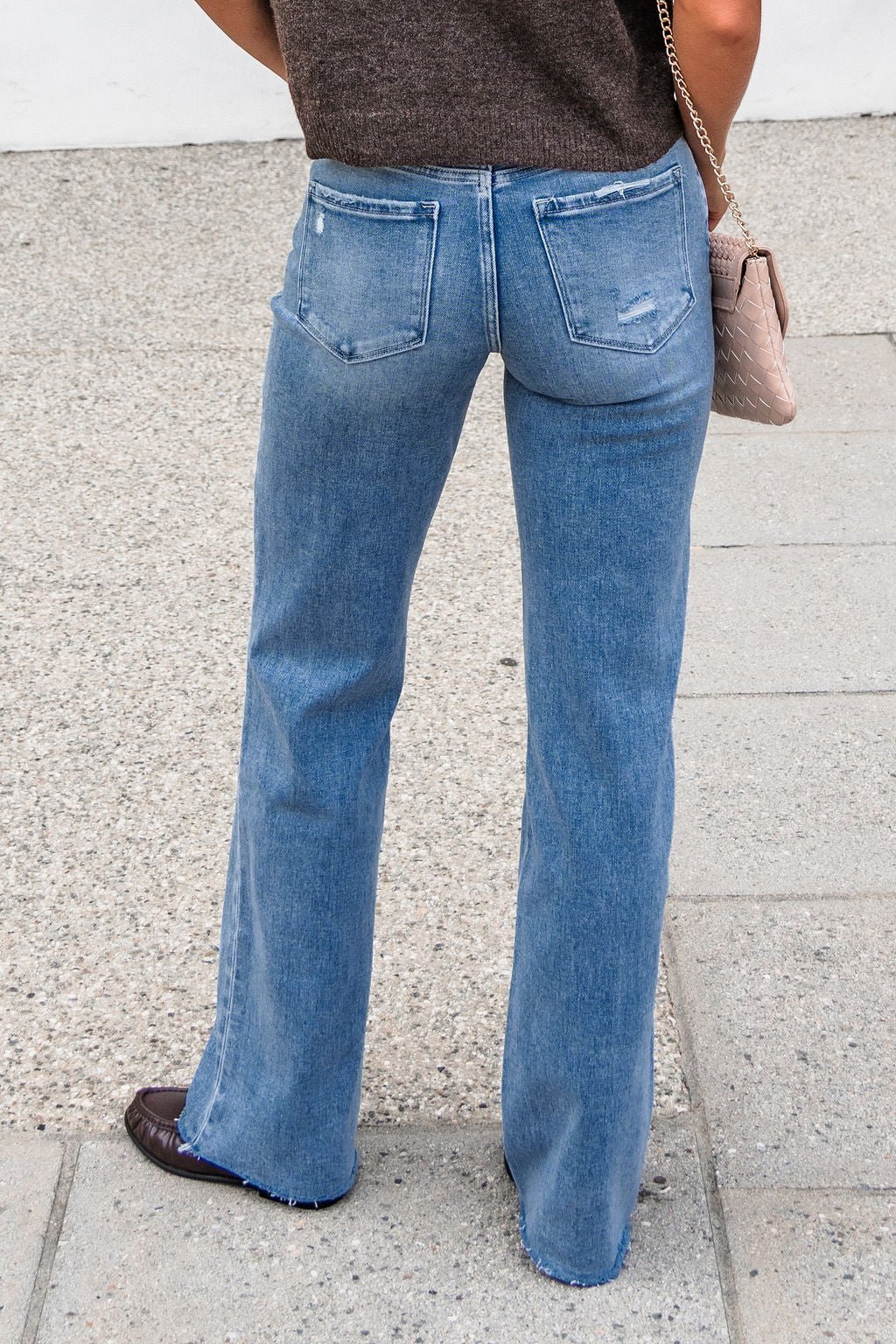 Person in Flying Monkey Distressed Wide Leg Jeans - Light Wash, brown shoes, and top stands on a sidewalk with a pink handbag over shoulder.