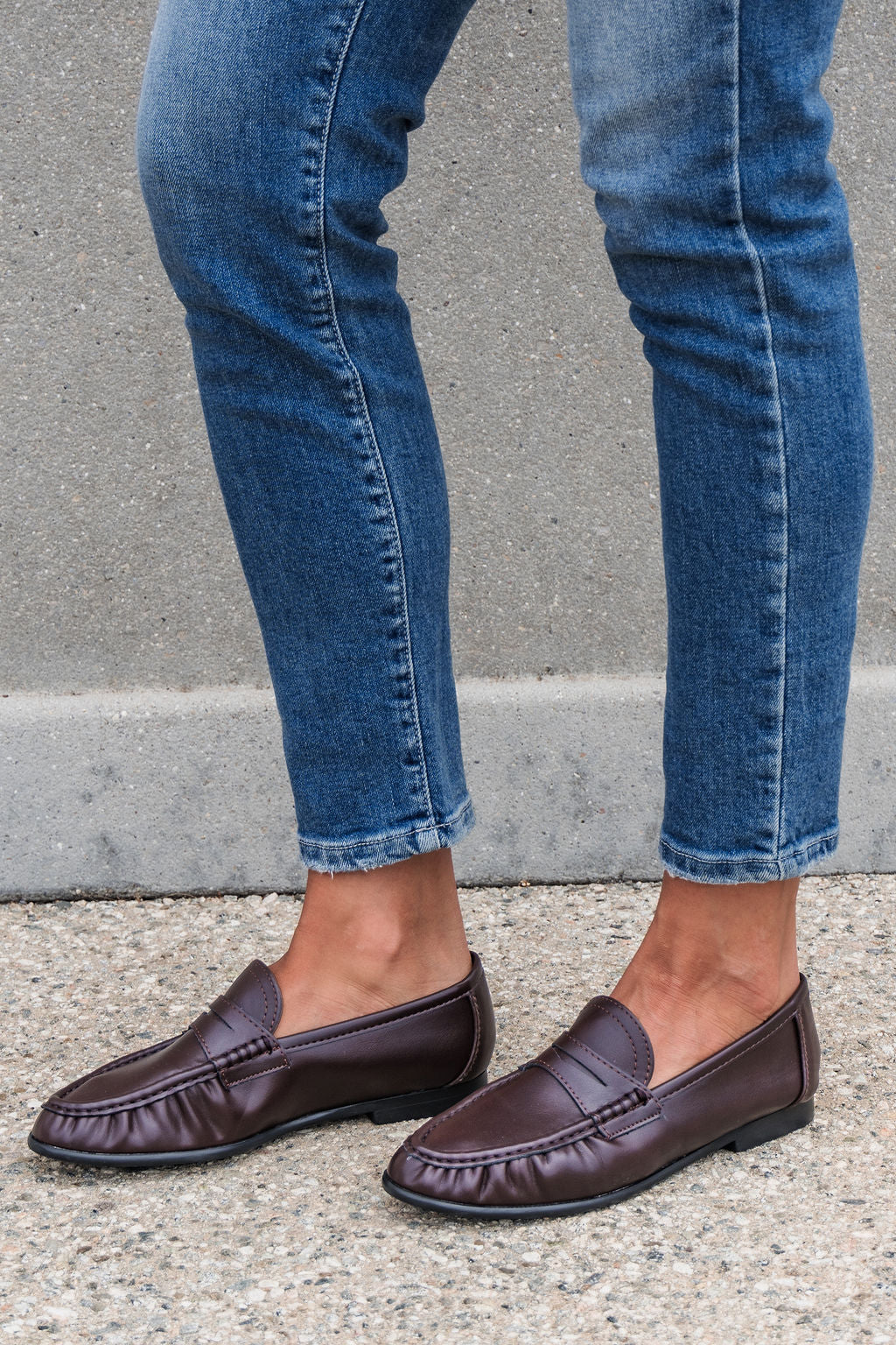 Person in blue jeans and Matisse Carlton Loafers - Brown stands on gravel before a concrete wall.