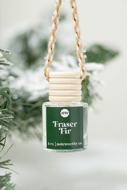 The Fraser Fir Car Diffuser hangs by a rope with a snowy pine branch behind, making it the perfect Christmas gift.