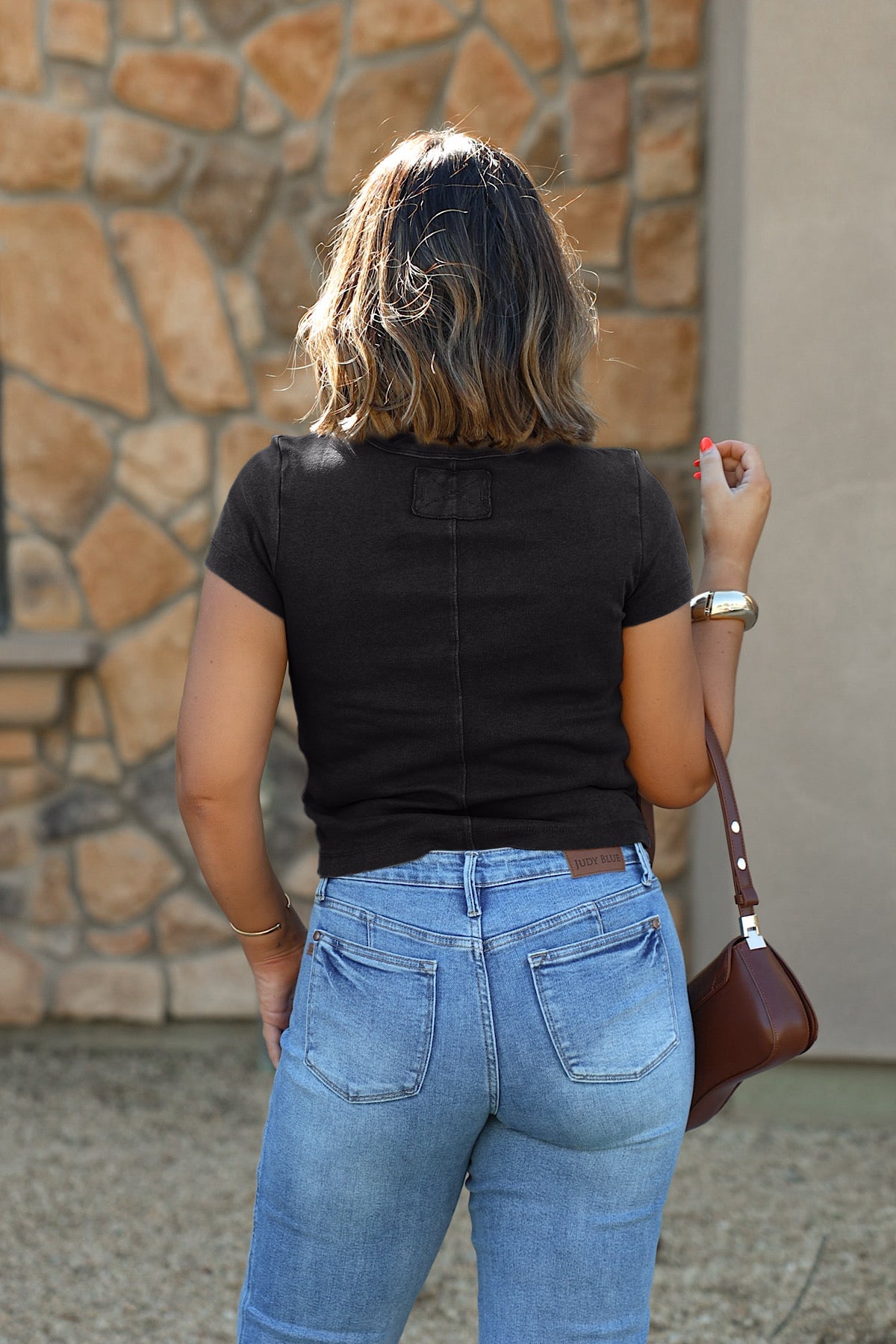 Woman with shoulder-length hair wears the Free People Atlas Crew Tee in Charcoal and blue jeans, holding a brown purse by a stone wall.