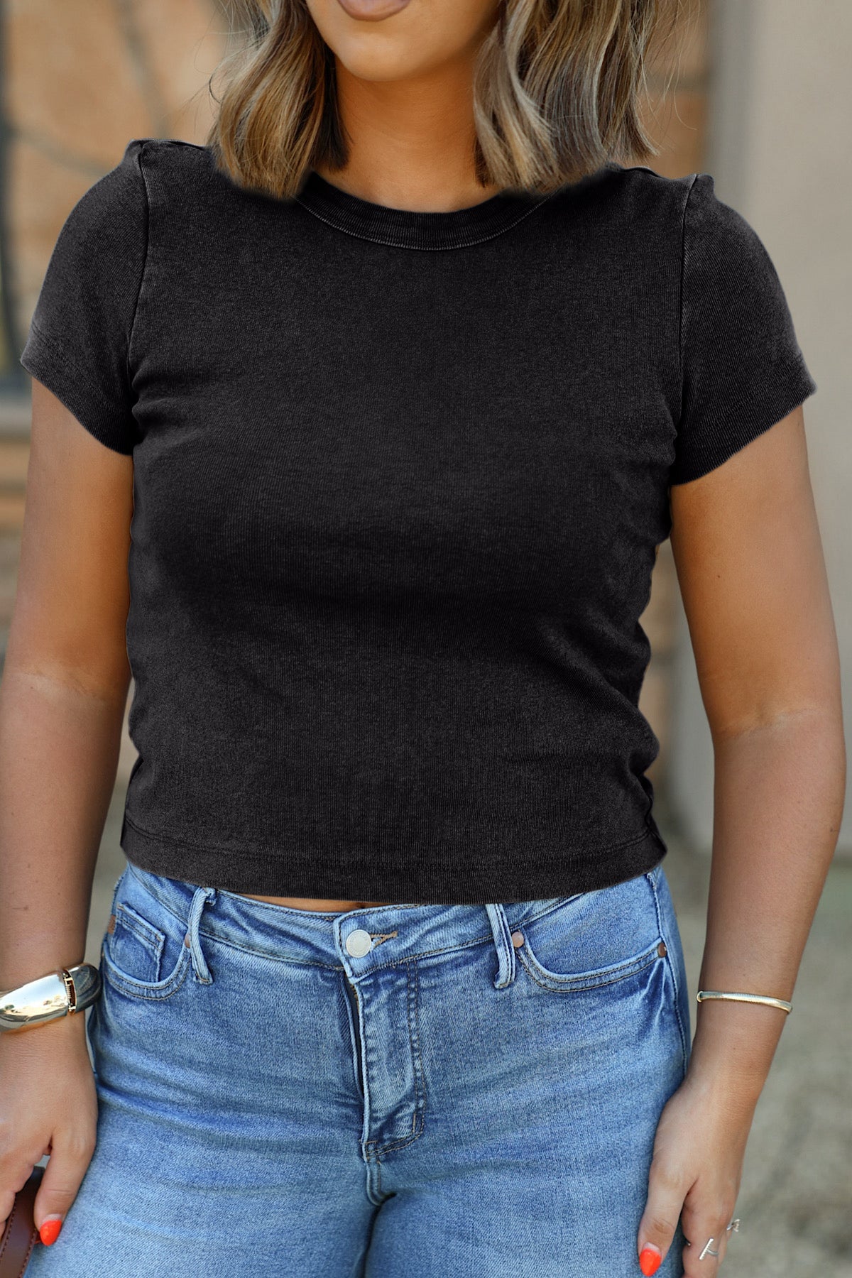 Woman wears a Free People Atlas Crew Tee in charcoal and blue jeans, standing outdoors with arms relaxed at her sides.