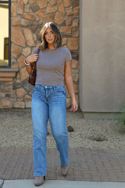Woman in a fitted Free People Atlas Crew Tee - Mocha and blue jeans walks outdoors on a brick path beside a stone wall.