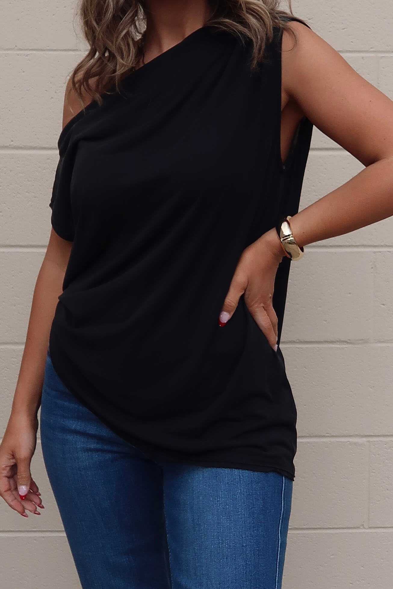 Woman in a Free People Black Off The Shoulder Cascadia Tunic and blue jeans, posing by a beige brick wall with one hand on her hip.
