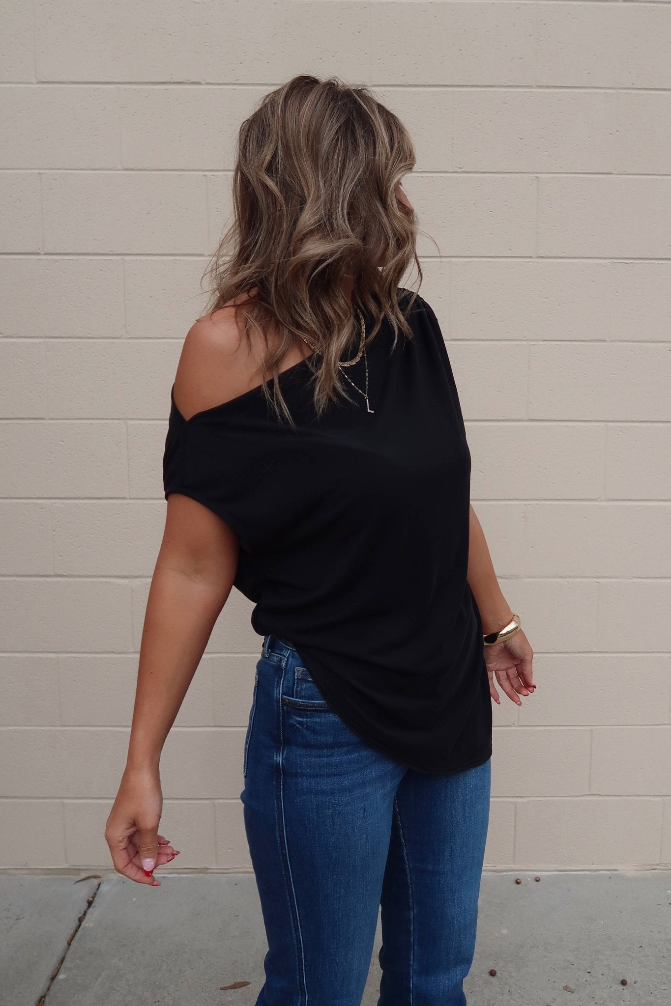 A woman with wavy hair wears a Free People Black Off The Shoulder Cascadia Tunic and blue jeans, standing by a beige brick wall.