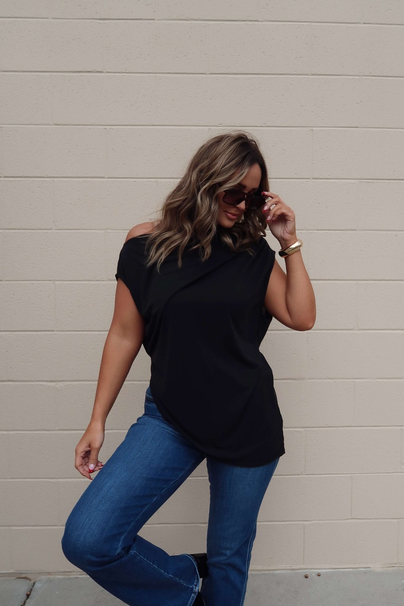 Woman in sunglasses, blue jeans, and a Free People Black Off The Shoulder Cascadia Tunic stands by a beige brick wall, smiling.