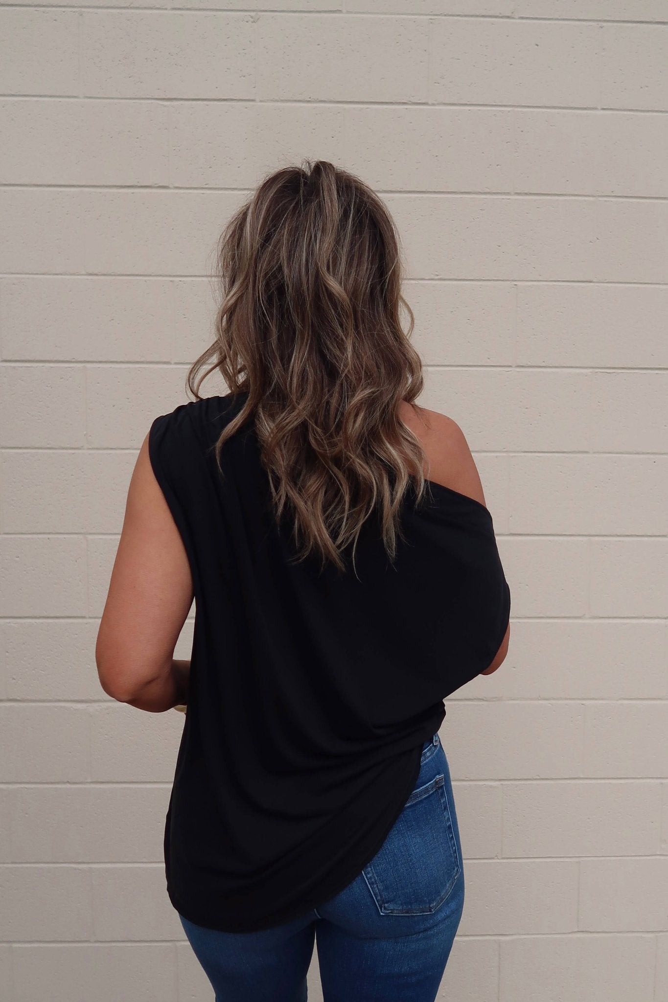 A woman with wavy brown hair in a Free People Black Off The Shoulder Cascadia Tunic and blue jeans stands by a beige brick wall.