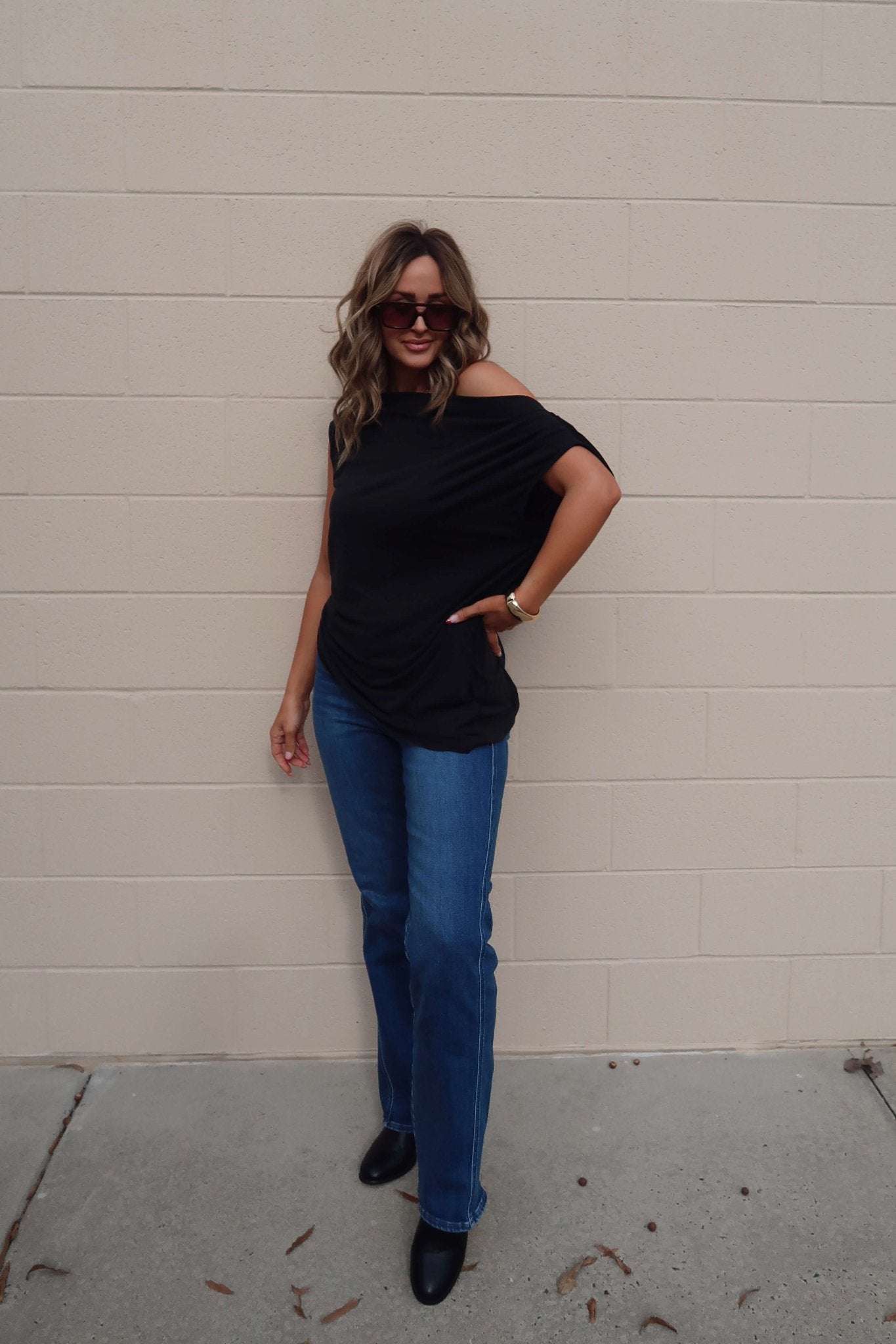 Woman in sunglasses, Free People Black Off The Shoulder Cascadia Tunic, and blue jeans poses casually by a beige brick wall.