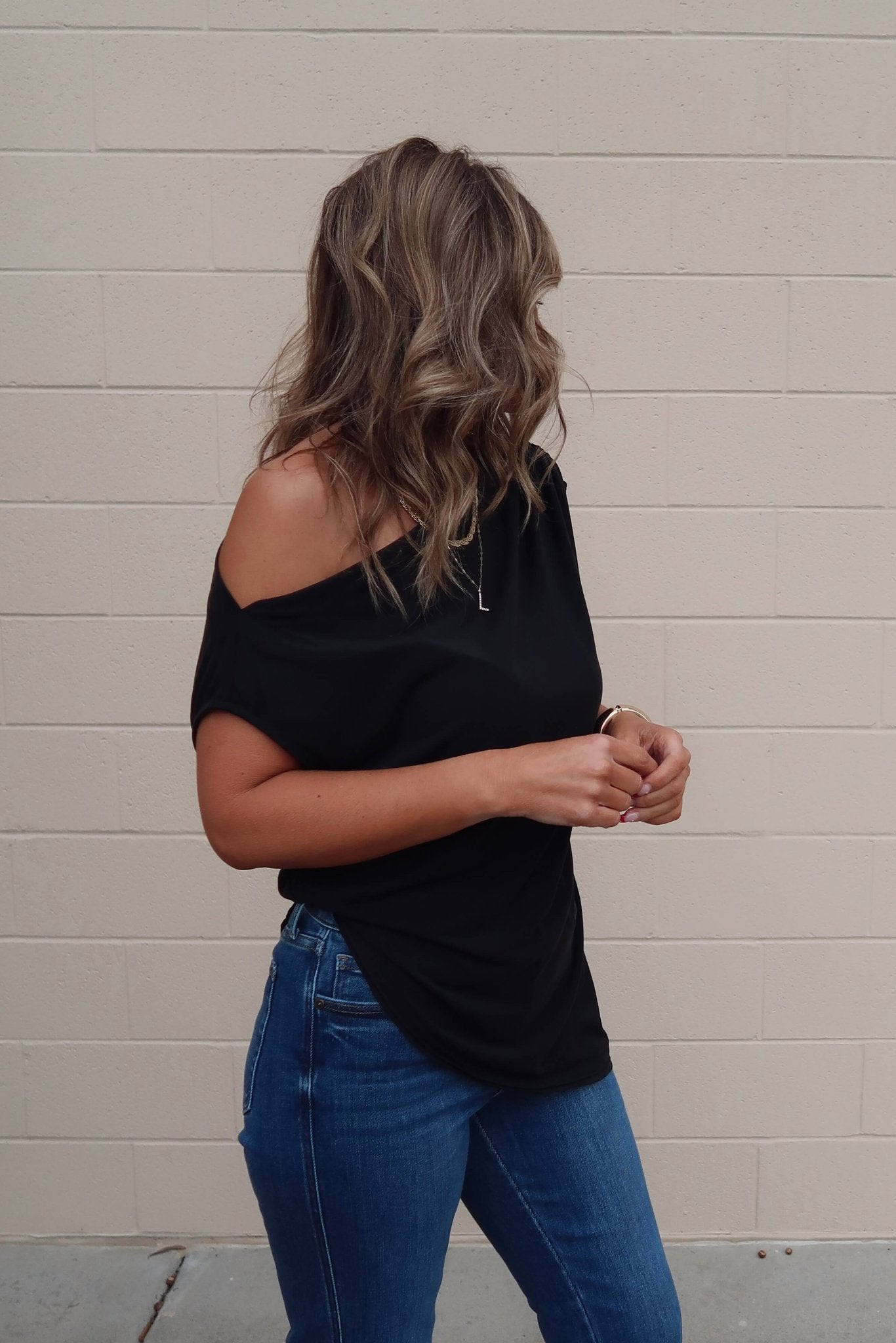 A woman in a Free People Black Off The Shoulder Cascadia Tunic and blue jeans stands by a beige brick wall, looking to the side.