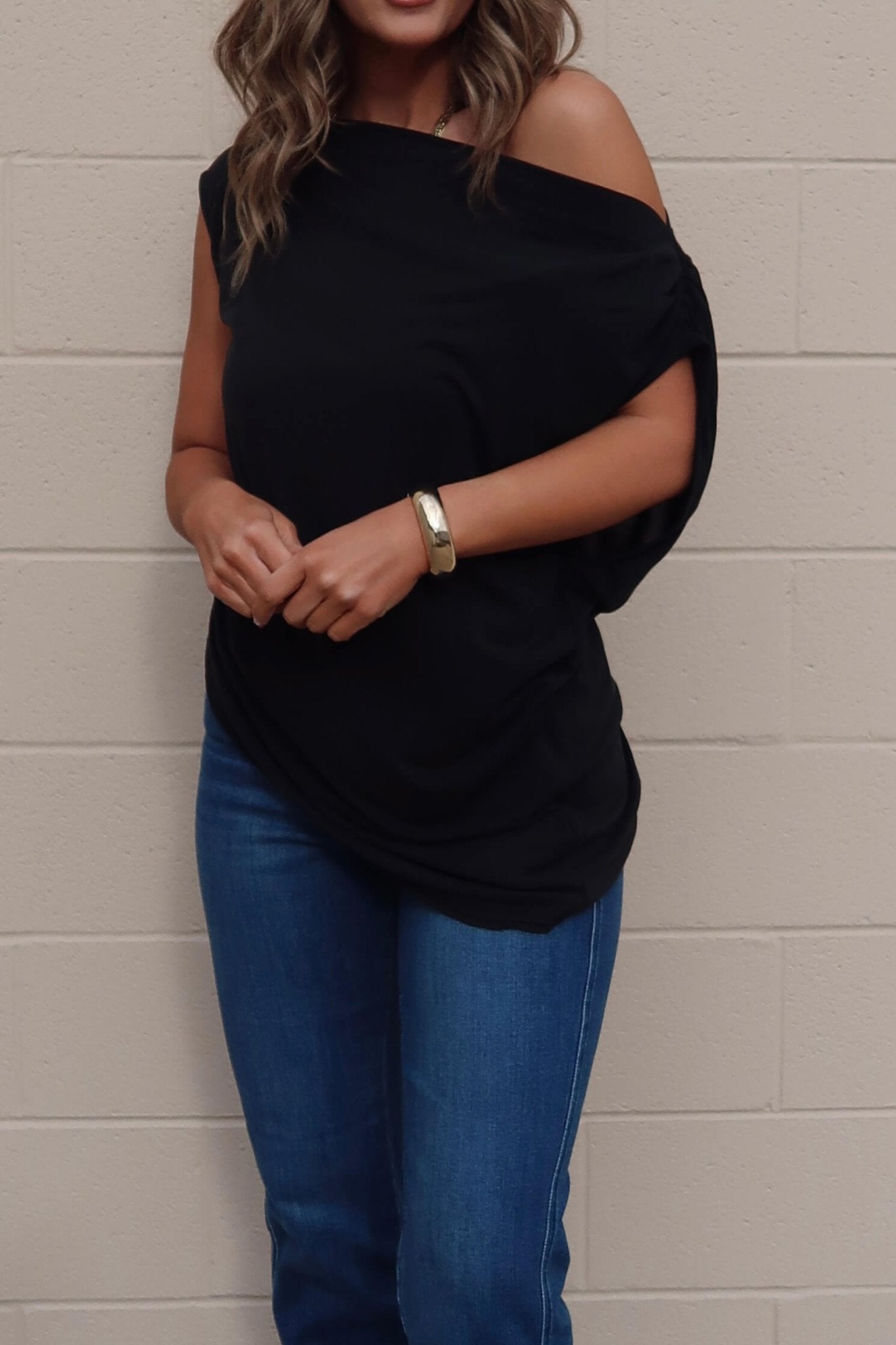 Woman in a Free People Black Off The Shoulder Cascadia Tunic, blue jeans, and gold bracelet by a light brick wall.