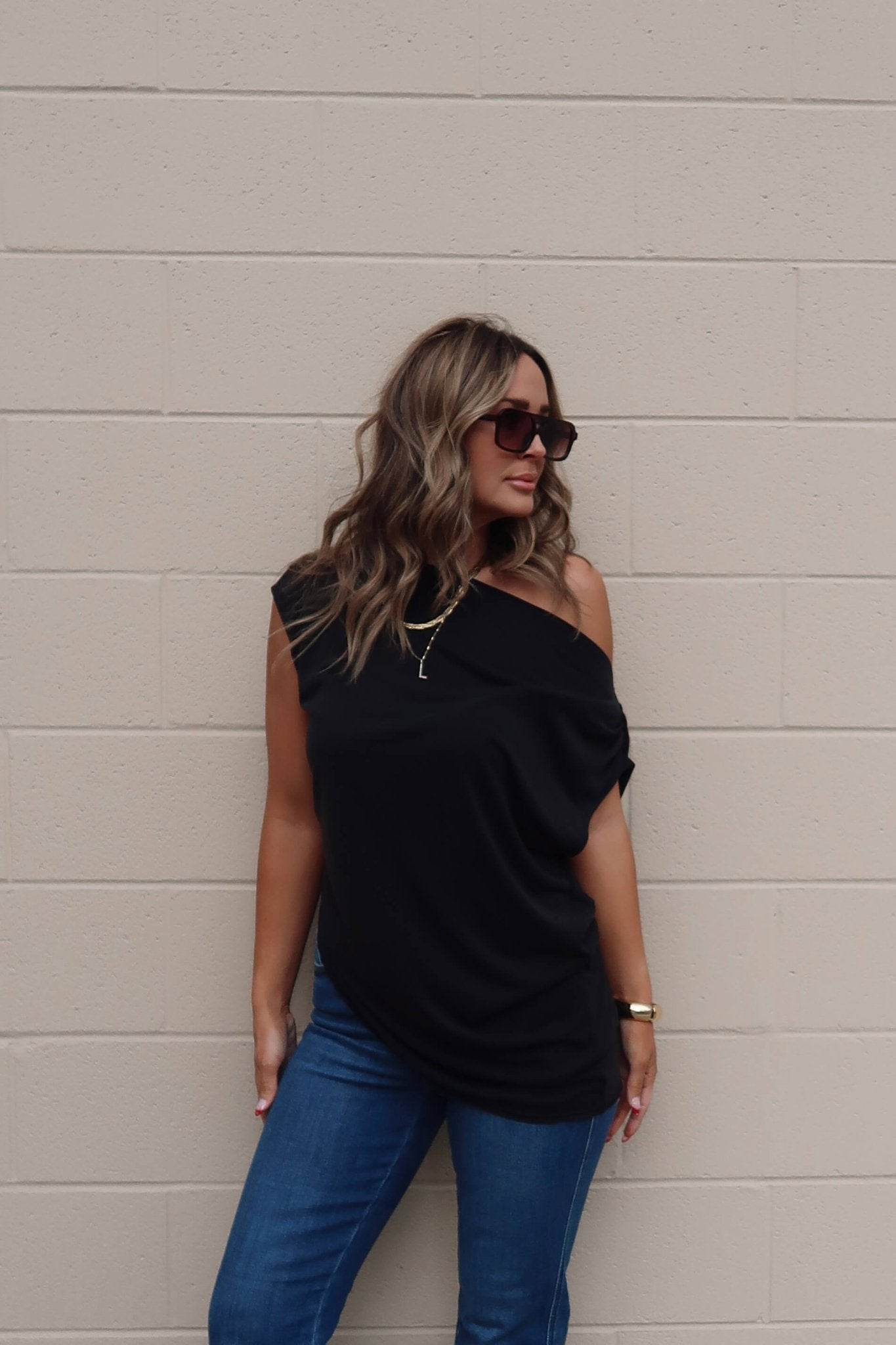 Woman with wavy hair in sunglasses and blue jeans wears a Free People Black Off The Shoulder Cascadia Tunic by a beige brick wall.