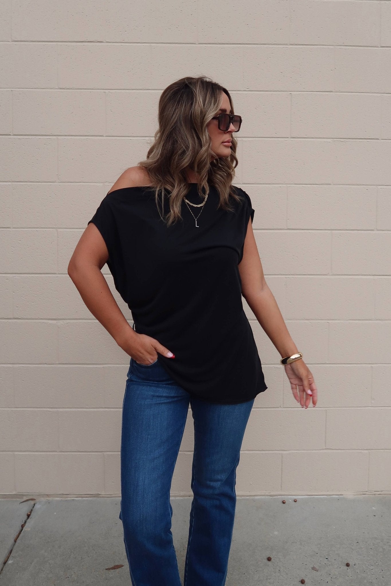 Woman with wavy hair wears a Free People Black Off The Shoulder Cascadia Tunic, blue jeans, and gold jewelry by a beige brick wall.