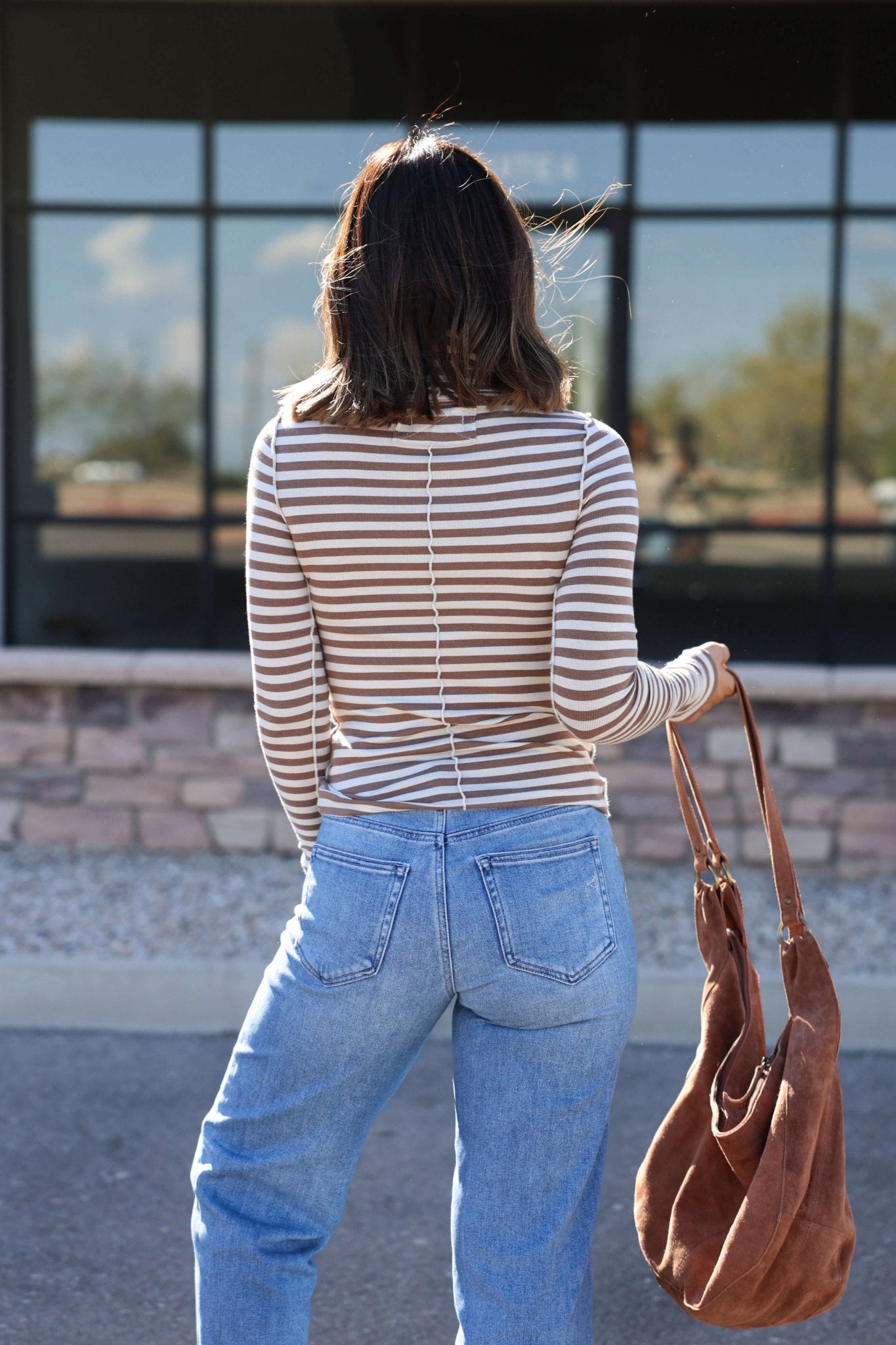 Woman with shoulder-length hair in the Free People Brown Good Good Striped Turtleneck Top and jeans, holding a brown bag, stands outside.