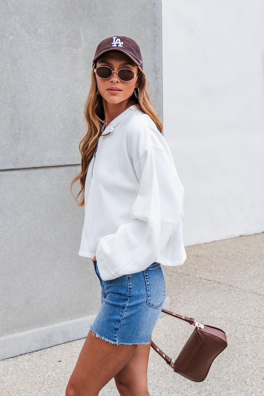 Woman in the Free People Celeste Quarter Zip in ivory, denim shorts, sunglasses, LA cap, and brown purse standing outdoors.
