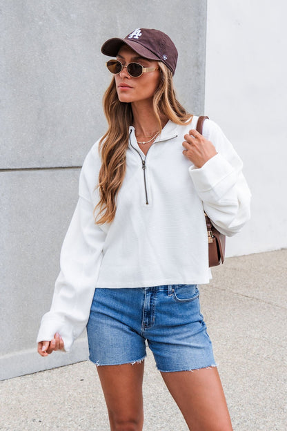 A woman outdoors in sunglasses, cap, and Free People Celeste Quarter Zip - Ivory pullover with denim shorts holds a bag strap.
