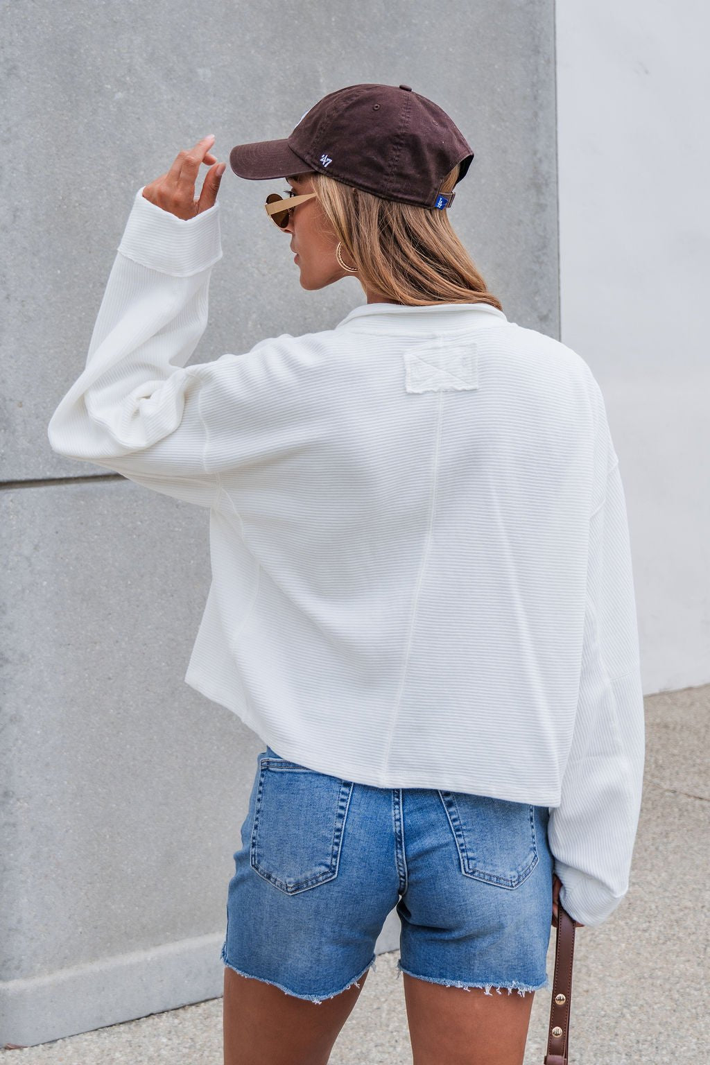 Woman wears a Free People Celeste Quarter Zip - Ivory, denim shorts, and a brown cap, standing outdoors with her back to the camera.
