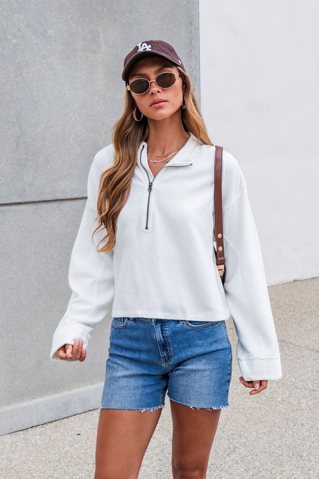 Woman in sunglasses wears the Free People Celeste Quarter Zip - Ivory, denim shorts, and a dark cap outdoors by a gray and white wall.