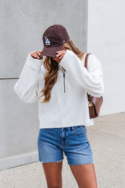 Person in a Free People Celeste Quarter Zip - Ivory, denim shorts, and LA cap adjusts their hat outside by a gray wall.