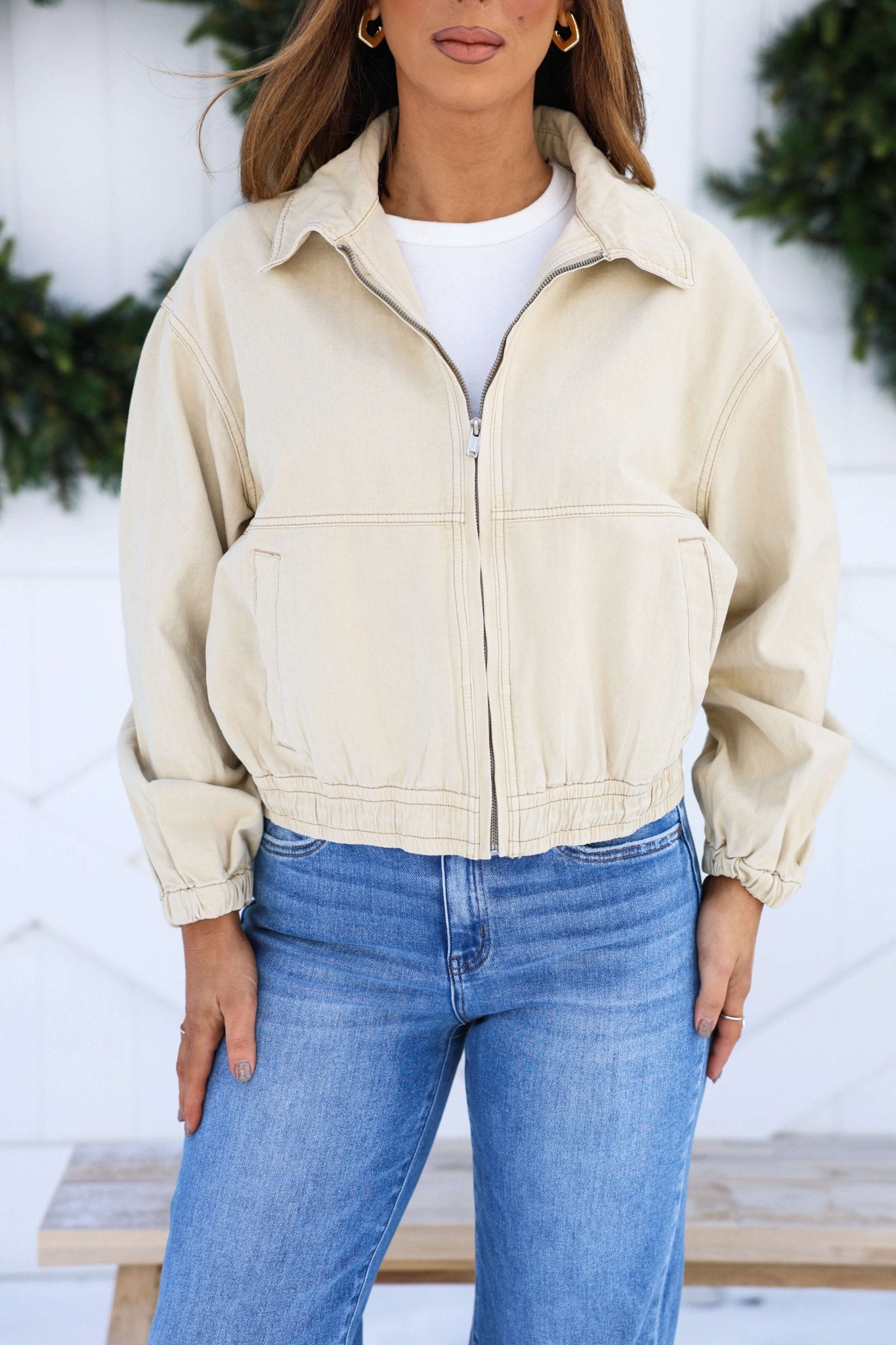 A woman in a Free People Cream Birdie Jacket over a white shirt and blue jeans stands outdoors before greenery.