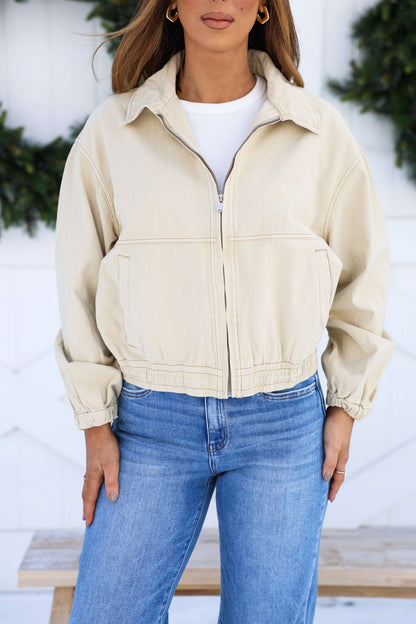 A woman in a Free People Cream Birdie Jacket over a white shirt and blue jeans stands outdoors before greenery.