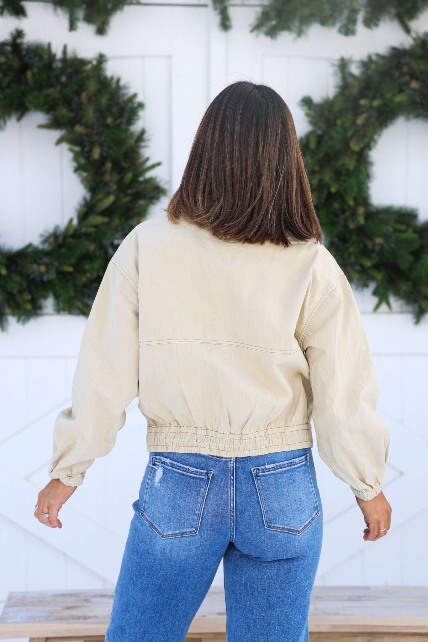 A woman in a Free People Cream Birdie Jacket and blue jeans stands outdoors, her shoulder-length hair turned away from the camera.
