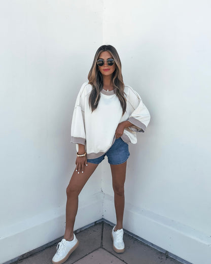 Woman in sunglasses, cream Free People Everything Pullover, and denim shorts poses by a white wall with one hand in her pocket.