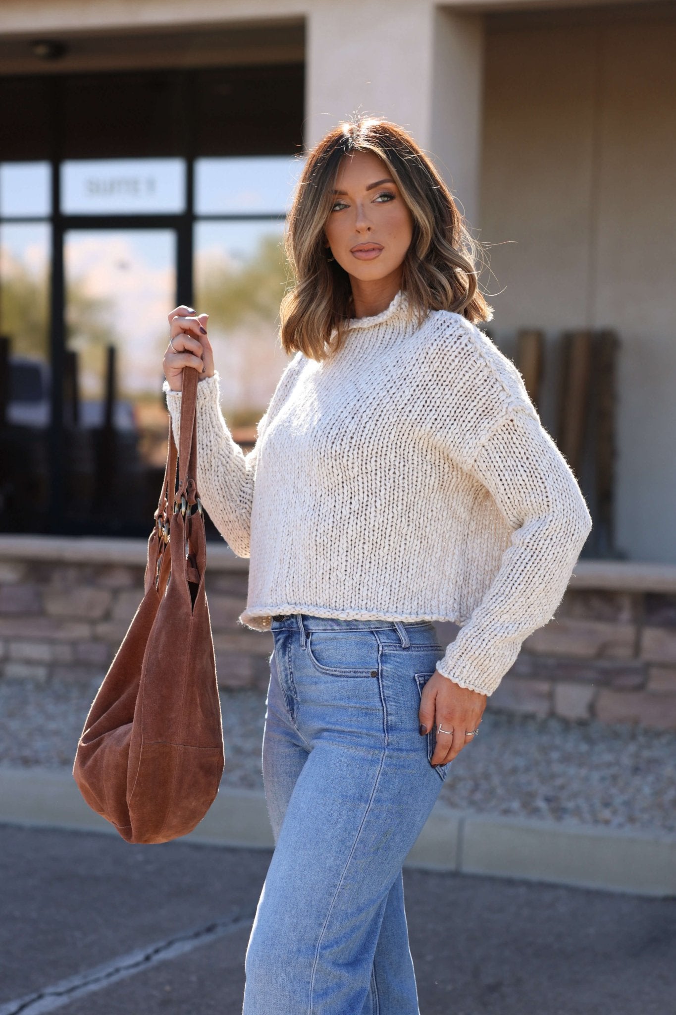 Woman in a cream Free People Opal Rollneck Sweater and jeans stands outside with a large brown bag—a cozy fall essential.