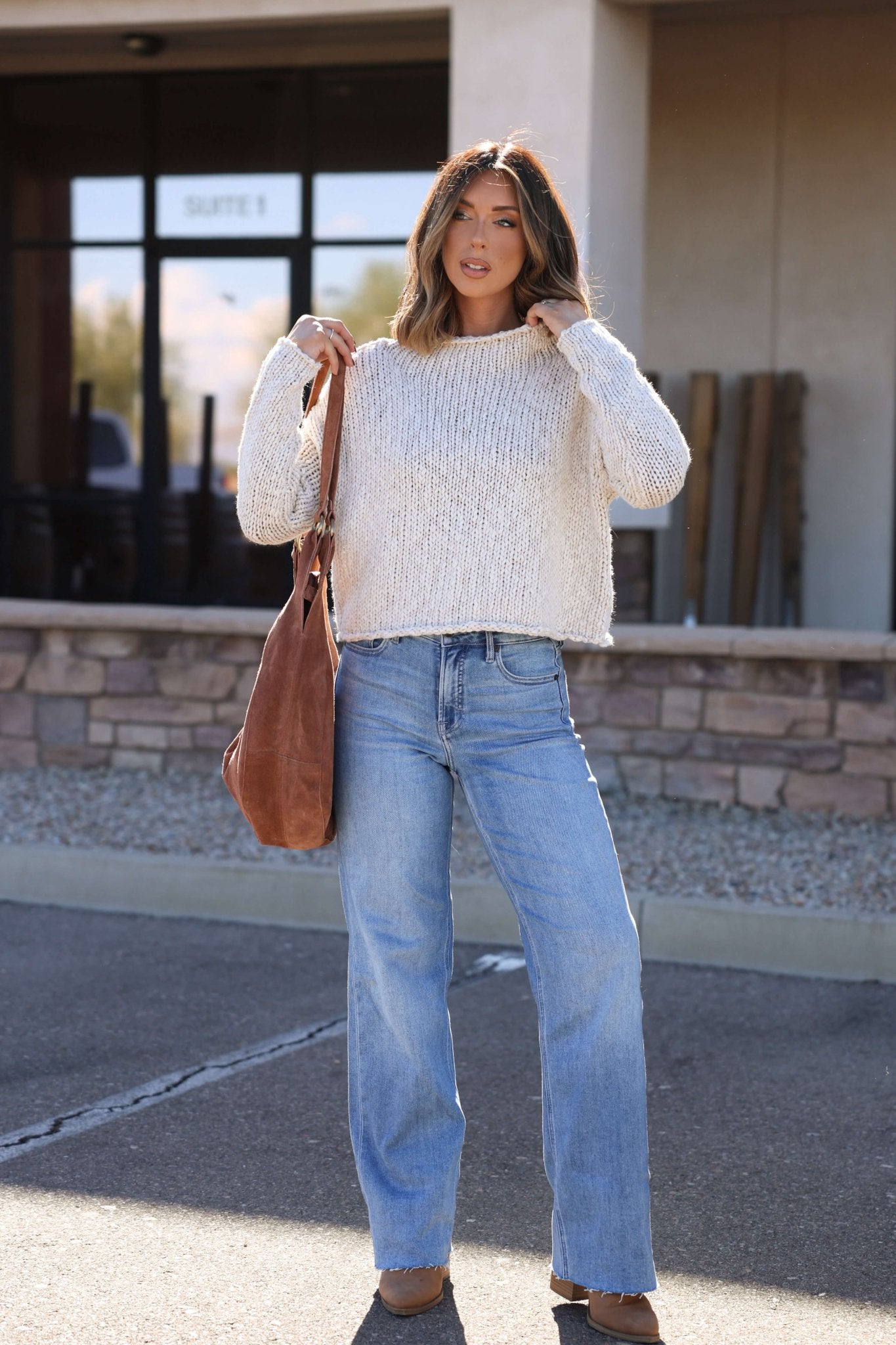 Woman standing outdoors in a cozy Free People Cream Opal Rollneck Sweater, blue jeans, brown shoes, and a large brown shoulder bag.