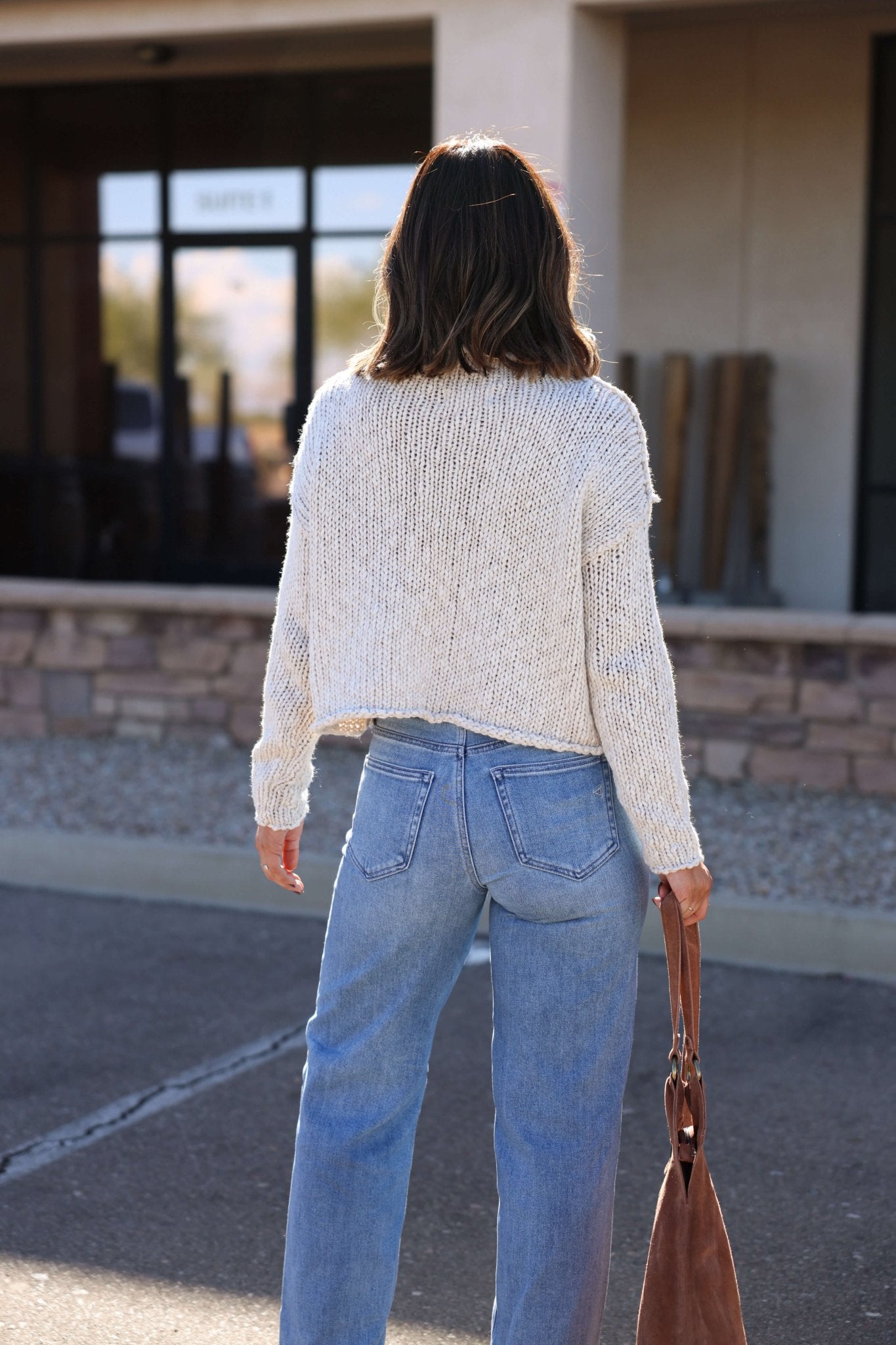 Woman with shoulder-length hair in a Free People Cream Opal Rollneck Sweater and jeans stands outside, holding a brown bag, back to camera.