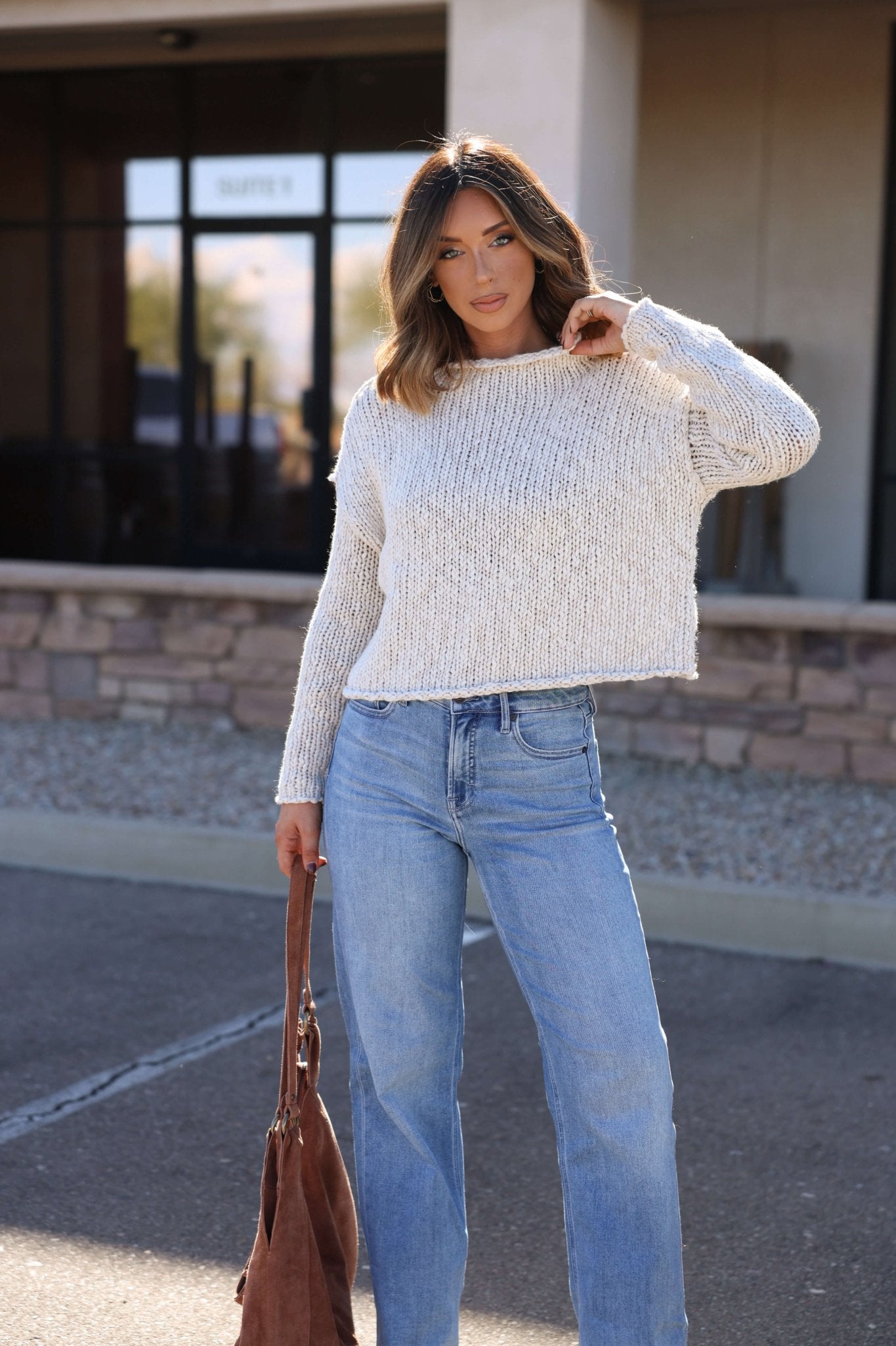 Woman in a Free People Cream Opal Rollneck Sweater and blue jeans stands outside, holding a brown bag with a building behind her.