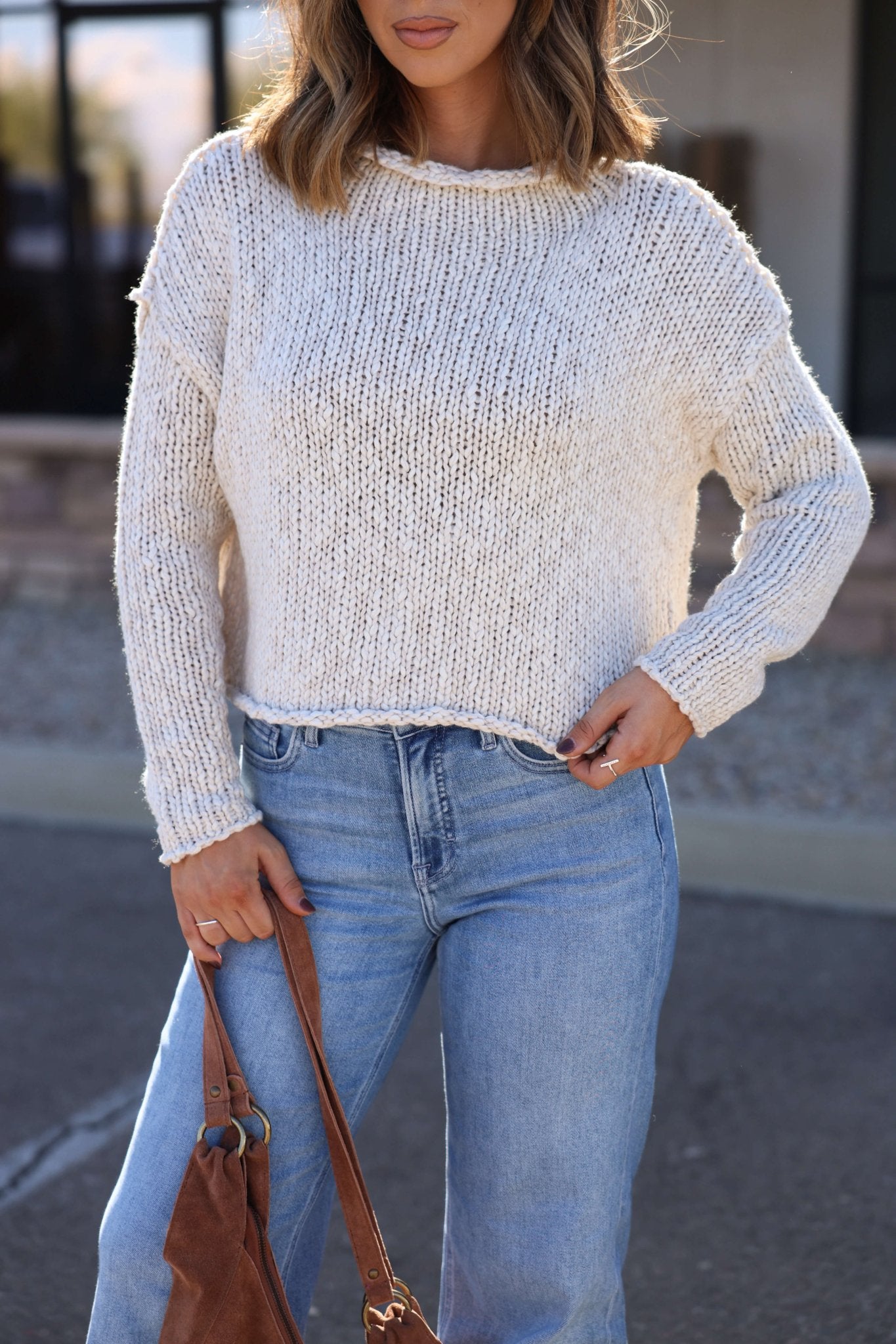 Woman in a Free People Cream Opal Rollneck Sweater and light blue jeans, holding a brown bag—perfect for your fall wardrobe outdoors.