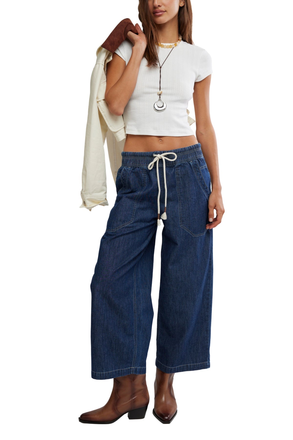 A woman in a white crop top, Free People Dark Blue Easy Peasy Pull On Jeans, brown boots, and necklaces holds a jacket over her shoulder.